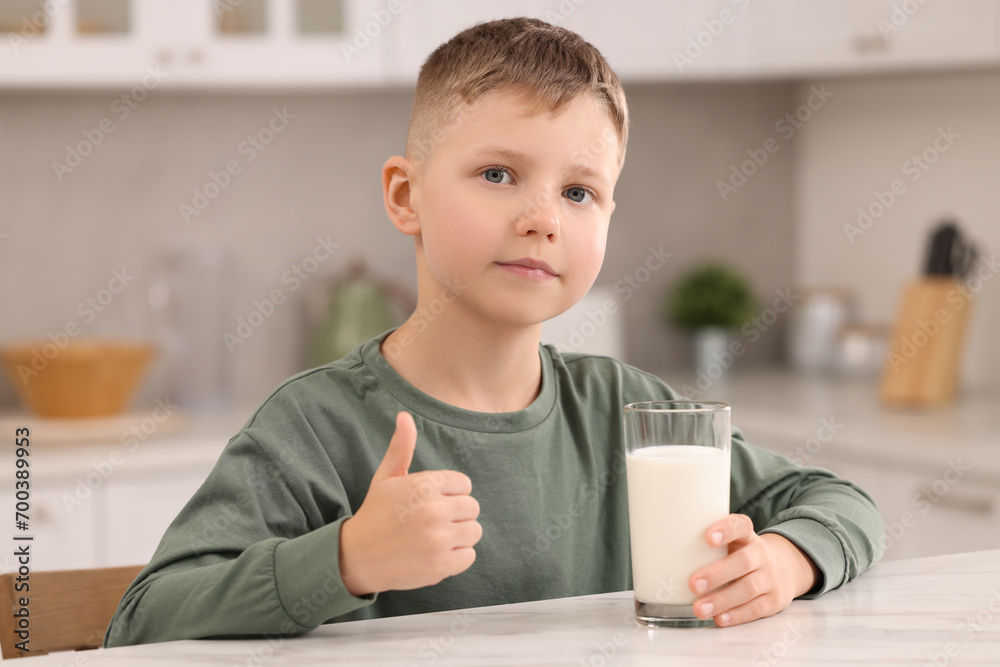 Cute boy with glass of fresh milk showing thumb up at white table in kitchen Cute boy with glass of fresh milk showing thumb up at white table in kitchen