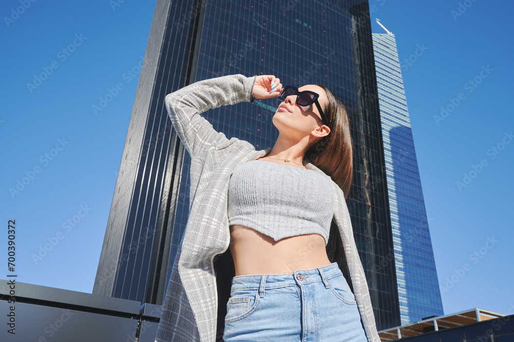 Dark haired woman with fit body wears sunglasses while posing artistically under the sunlight between modern buildings. She wears a top, blazer and jeans. Dark haired woman with fit body wears sunglasses while posing artistically under the sunlight between modern buildings. She wears a top, blazer and jeans.
