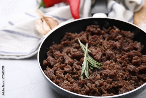 Fried ground meat in frying pan and rosemary on white tiled table, closeup