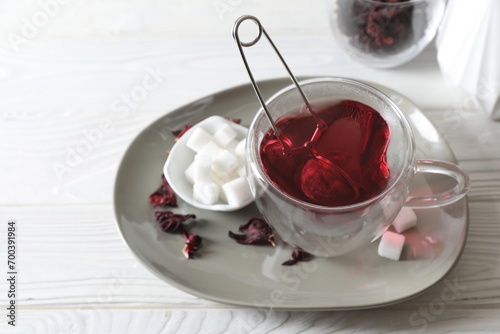 Delicious hibiscus tea in cup, sugar cubes and dry roselle petals on white wooden table, above view. Space for text