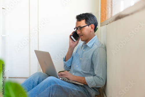 The young man asian is working from home in the living room, using his mobile phone and computer to command his work remotely.