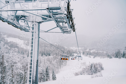Chairlift pole with chairs moving on cables uphill above a snowy forest