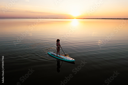 A woman in mohawk shorts stands on a SUP board at sunset in a lake against a pink-blue sky and water. Horizontal photo © diy13