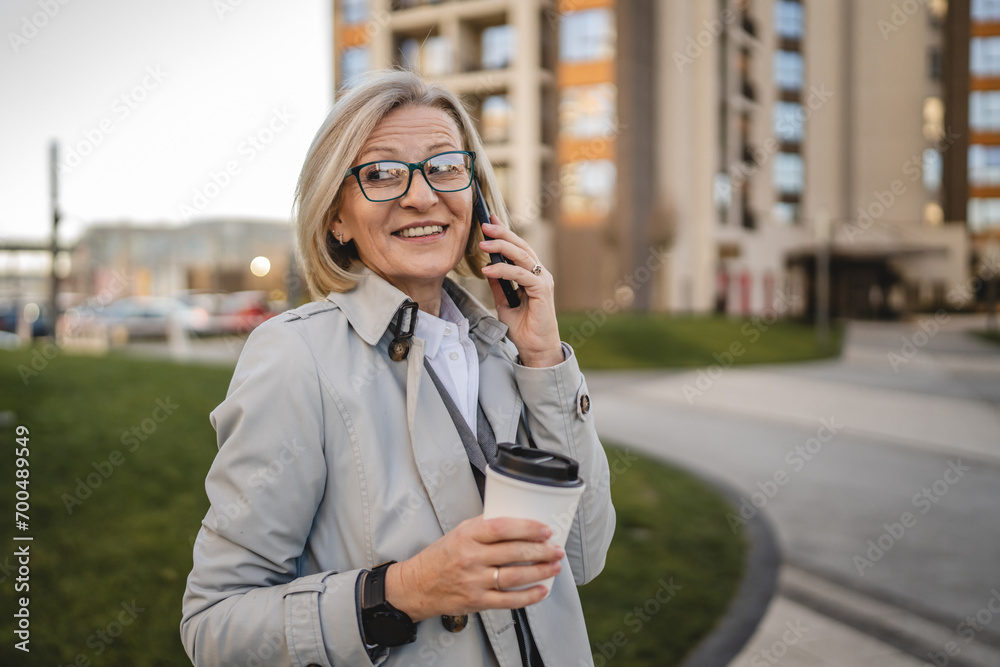 One mature woman outdoors in front of buildings talking on mobile phone and holding a to-go coffee, modern business and lifestyle concept One mature woman outdoors in front of buildings talking on mobile phone and holding a to-go coffee, modern business and lifestyle concept