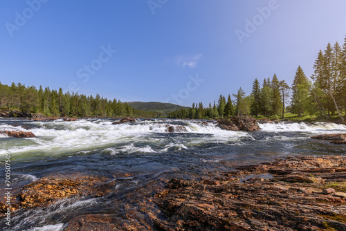 A dynamic summer view of the cascading Namsen River in Namsskogan, Trondelag, Norway, with large rocks and solitary trees in its course, framed by rugged stones of its rocky banks