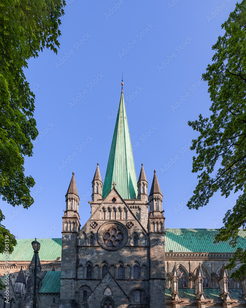 The main entrance of Nidaros Cathedral (Nidarosdomen) in Trondheim, Norway, is majestically framed by green trees on either side, highlighted by its striking pointed spire The main entrance of Nidaros Cathedral (Nidarosdomen) in Trondheim, Norway, is majestically framed by green trees on either side, highlighted by its striking pointed spire