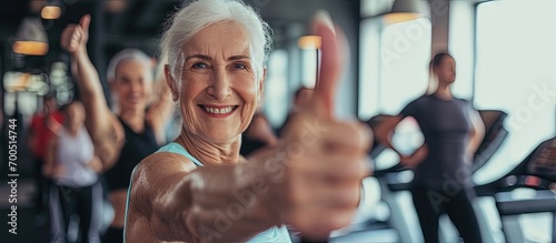 Cheerful senior woman gesturing thumbs up with people exercising in the background at fitness studio. Creative Banner. Copyspace image photo