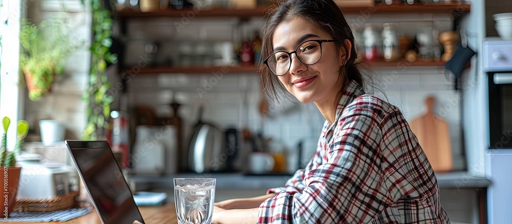 A young woman of oriental origin in a hijab is sitting at home in the kitchen at the table with a laptop and holding a glass of pure mineral water Leads a healthy lifestyle smiles advertises