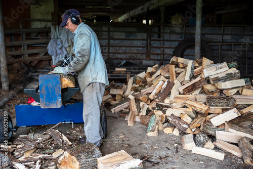 Man with work gloves safety ear protectors uses hydraulic log splitter to cut cordwood with woodpile of split firewood logs behind him