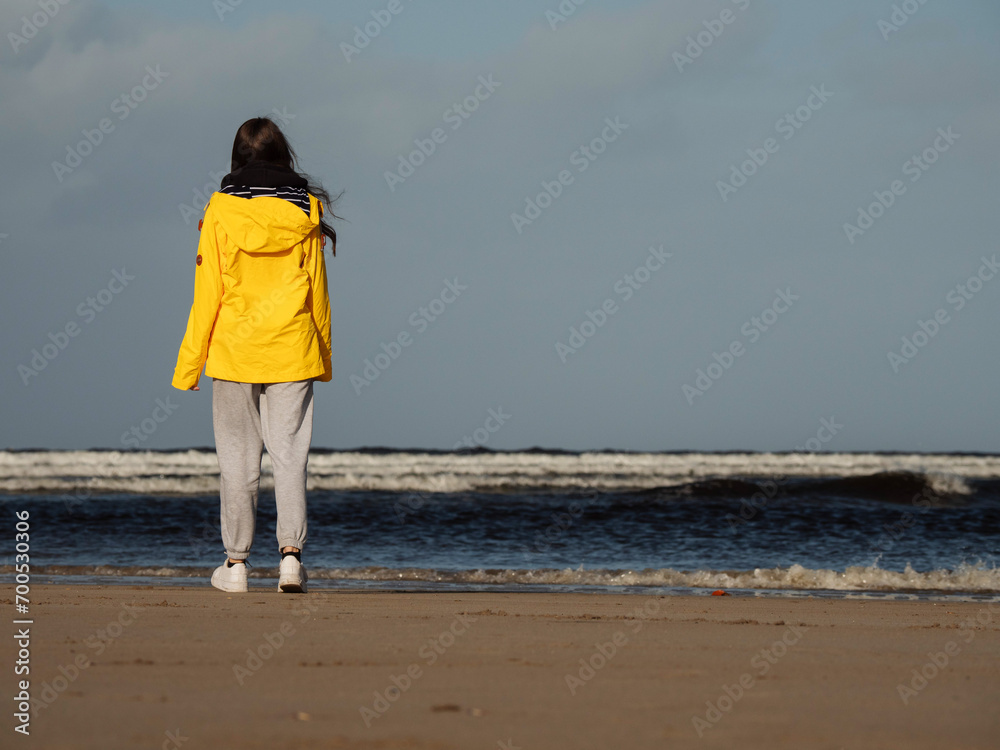 Teenager girl walking on a beach back to viewer. Young model in yellow jacket enjoying outdoor activity on fresh air. Selective focus. Cloudy sky. Travel and tourism concept.