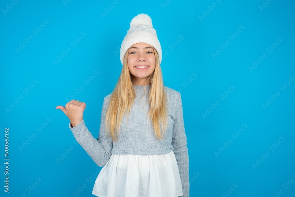 Lovely Caucasian blonde girl wearing blue sweater and wool beanie pointing aside with forefinger, showing at copy space having news about bargains Lovely Caucasian blonde girl wearing blue sweater and wool beanie pointing aside with forefinger, showing at copy space having news about bargains