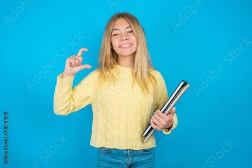 Caucasian blonde girl wearing yellow sweater holding notebooks smiling and confident gesturing with hand doing small size sign with fingers looking and the camera. Measure concept