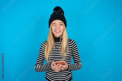 Excited beautiful caucasian teen girl wearing striped sweater winking and eye hold smart phone use read social network news