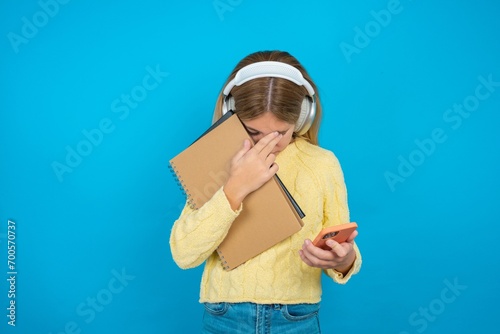 beautiful caucasian teen girl wearing yellow sweater  looking at smart phone feeling sad holding hand on face.