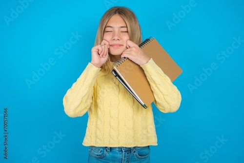Pleased beautiful caucasian teen girl wearing yellow sweater with closed eyes keeps hands near cheeks and smiles tenderly imagines something very pleasant