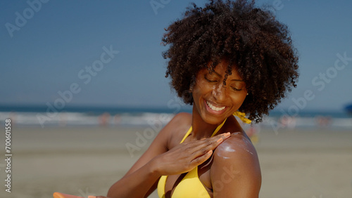 Close up of a happy smiling young black woman is applying a sunscreen or sun tanning lotion on a shoulder to take care of her skin on a seaside beach during holidays vacation.
