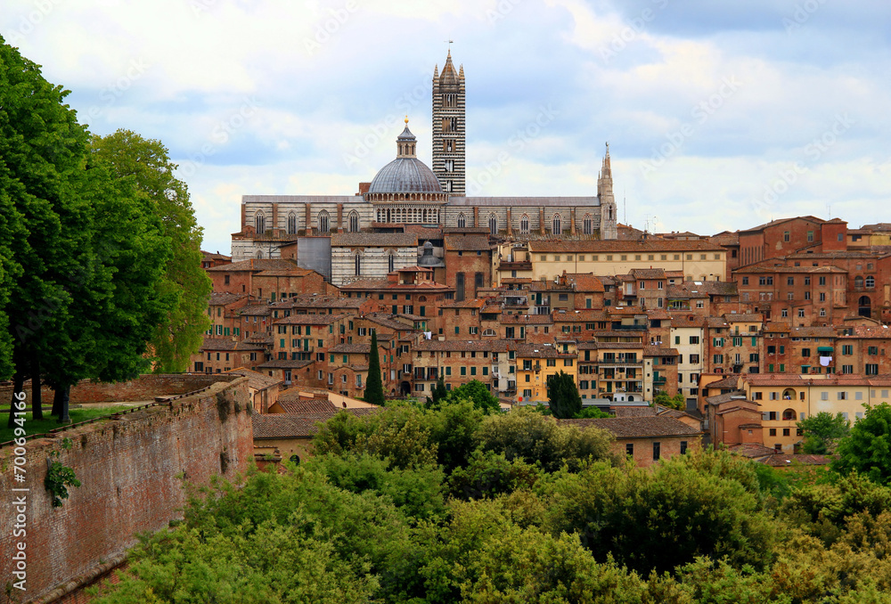 Panoramic view of the historic part of the city of Siena with the Duomo di Siena and green trees and bushes in the foreground against a blue sky with clouds in the Tuscany region of Italy