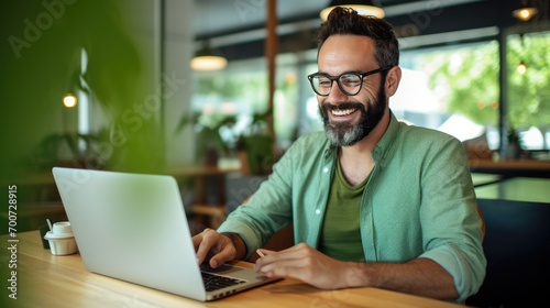 Cheerful man with a beard and glasses working on a laptop at a wooden desk photo