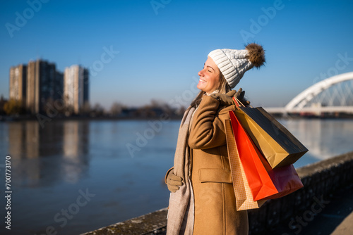 Happy woman in warm clothing with shopping bags enjoys standing by river on sunny winter day. 