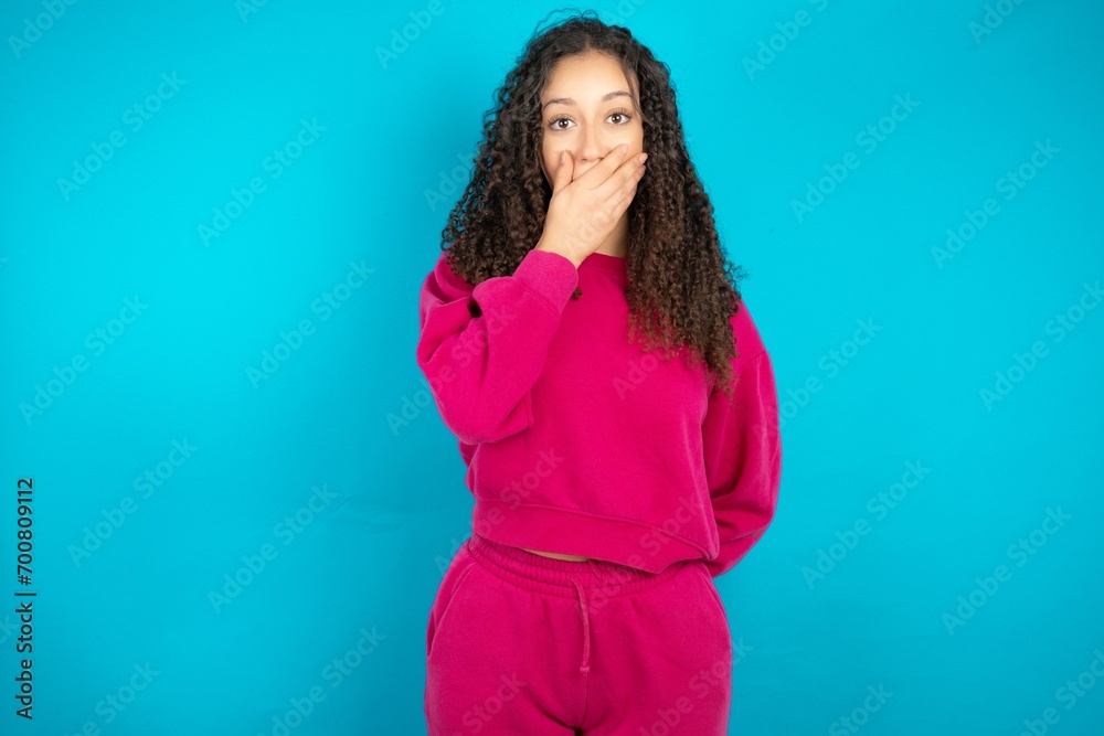 Emotional Beautiful young girl wearing pink tracksuit on blue background gasps from astonishment, covers opened mouth with palm, looks shocked at camera.
