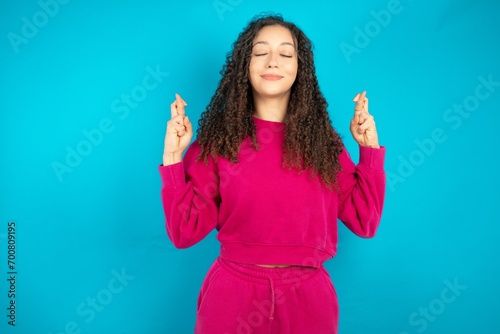 Beautiful young girl wearing pink tracksuit on blue background has big hope, crosses fingers, believes in good fortune, smiles broadly. People and wish concept
