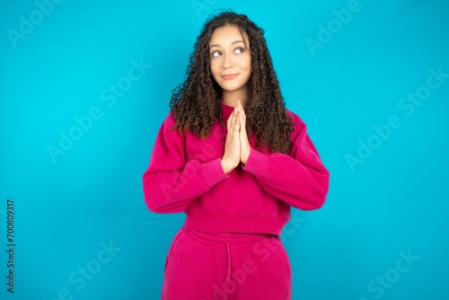 Charming cheerful Beautiful young girl wearing pink tracksuit on blue background making up plan in mind holding hands together, setting up an idea. photo
