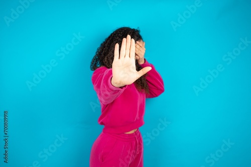 Beautiful young girl wearing pink tracksuit on blue background covers eyes with palm and doing stop gesture, tries to hide from everybody.