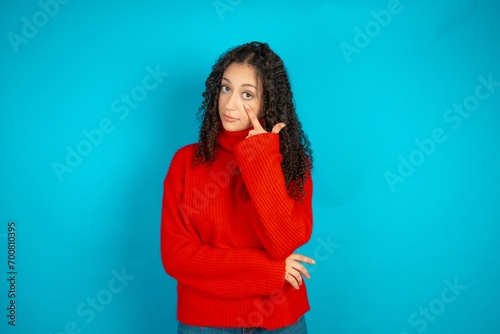 Beautiful teen girl wearing knitted red sweater over blue background Pointing to the eye watching you gesture, suspicious expression.