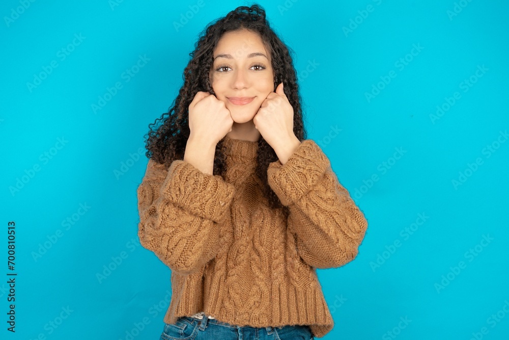 Happy beautiful teen girl wearing brown knitted sweater keeps fists on cheeks smiles broadly and has positive expression being in good mood Happy beautiful teen girl wearing brown knitted sweater keeps fists on cheeks smiles broadly and has positive expression being in good mood