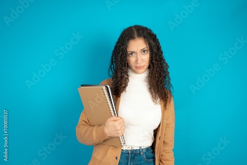 Portrait of dissatisfied beautiful teen woman wearing beige knitted jacket smirks face, purses lips and looks with annoyance at camera, discontent hearing something unpleasant