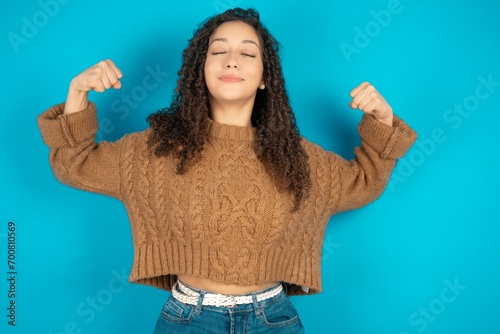 Strong powerful beautiful teen girl wearing brown knitted sweater toothy smile, raises arms and shows biceps. Look at my muscles!