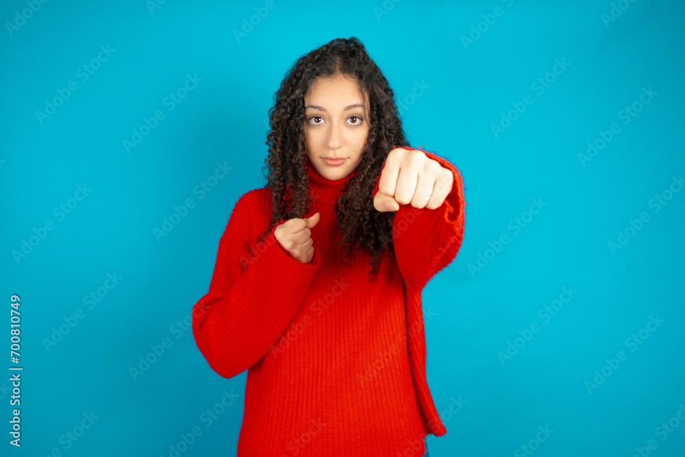 Beautiful teen girl wearing knitted red sweater over blue background Punching fist to fight, aggressive and angry attack, threat and violence Beautiful teen girl wearing knitted red sweater over blue background Punching fist to fight, aggressive and angry attack, threat and violence