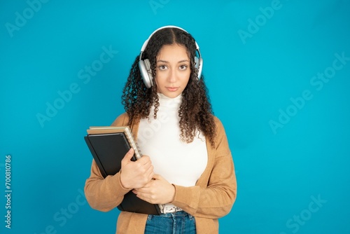 Serious displeased beautiful teen woman wearing beige knitted jacket looks puzzled at camera being angry wears stereo headphones listens music while walking at street photo
