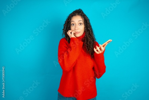 Afraid funny beautiful teen girl wearing red knitted sweater holding telephone and bitting nails photo