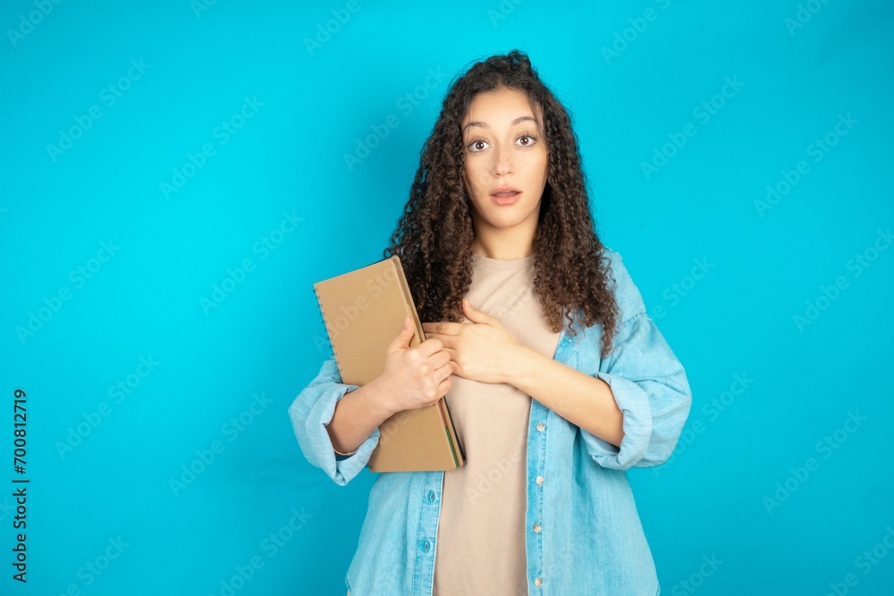 Scared beautiful arab student carries notebooks looks with frightened expression, keeps hands on chest, being puzzled to notice something strange, People, hush reaction and emotions. Scared beautiful arab student carries notebooks looks with frightened expression, keeps hands on chest, being puzzled to notice something strange, People, hush reaction and emotions.