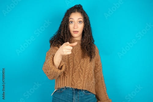 Shocked Beautiful teen girl wearing knitted sweater over blue background points at you with stunned expression