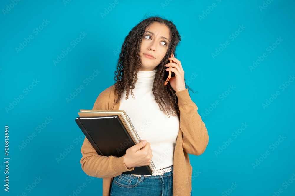 beautiful arab student carries notebooks speaks on mobile phone spends free time indoors calls to friend. beautiful arab student carries notebooks speaks on mobile phone spends free time indoors calls to friend.
