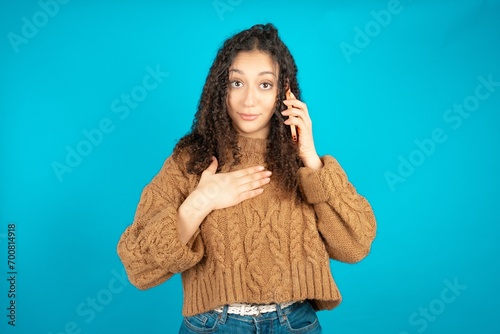 Smiling Beautiful teen girl wearing knitted sweater over blue background talks via cellphone, enjoys pleasant great conversation. People, technology, communication concept