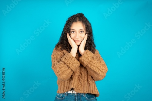 Happy Beautiful teen girl wearing knitted sweater over blue background touches both cheeks gently, has tender smile, shows white teeth, gazes positively straightly at camera,