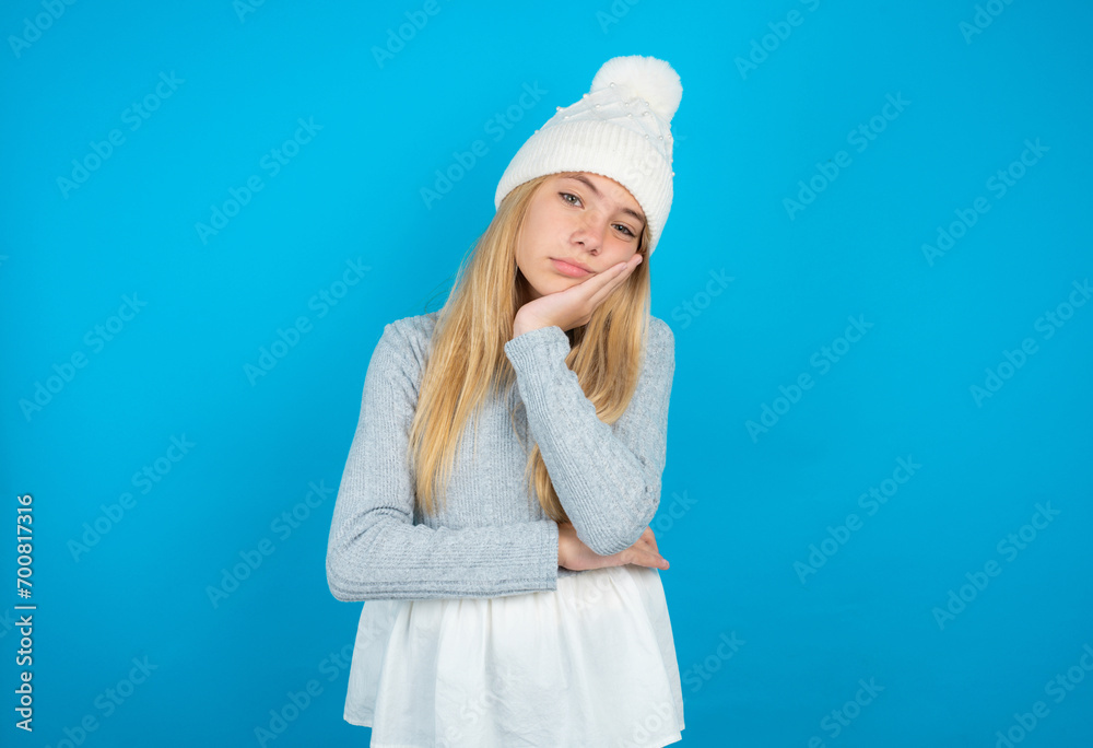 Very bored Teen caucasian girl wearing blue knitted sweater and woolly hat holding hand on cheek while support it with another crossed hand, looking tired and sick,
