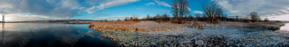 Panorama of the sunrise over the winter river. The sun rises over the lake. Autumn frost. The frozen river is covered with thin ice, the grass glistens with frost.