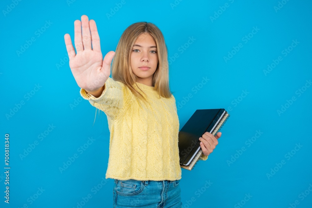 Beautiful kid girl wearing yellow sweater holding notebook doing stop sing with palm of the hand. Warning expression with negative and serious gesture on the face. Beautiful kid girl wearing yellow sweater holding notebook doing stop sing with palm of the hand. Warning expression with negative and serious gesture on the face.
