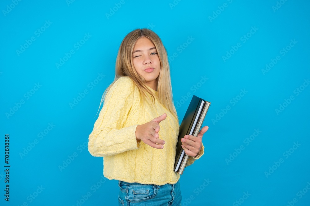 Joyful Beautiful kid girl wearing yellow sweater holding notebook wink and points index fingers at camera, chooses someone, makes finger gun pistol. Joyful Beautiful kid girl wearing yellow sweater holding notebook wink and points index fingers at camera, chooses someone, makes finger gun pistol.