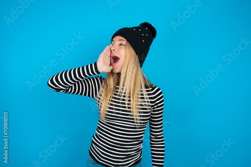 Teen caucasian girl wearing striped sweater and woolly hat shouting and screaming loud to side with hand on mouth. Communication concept.