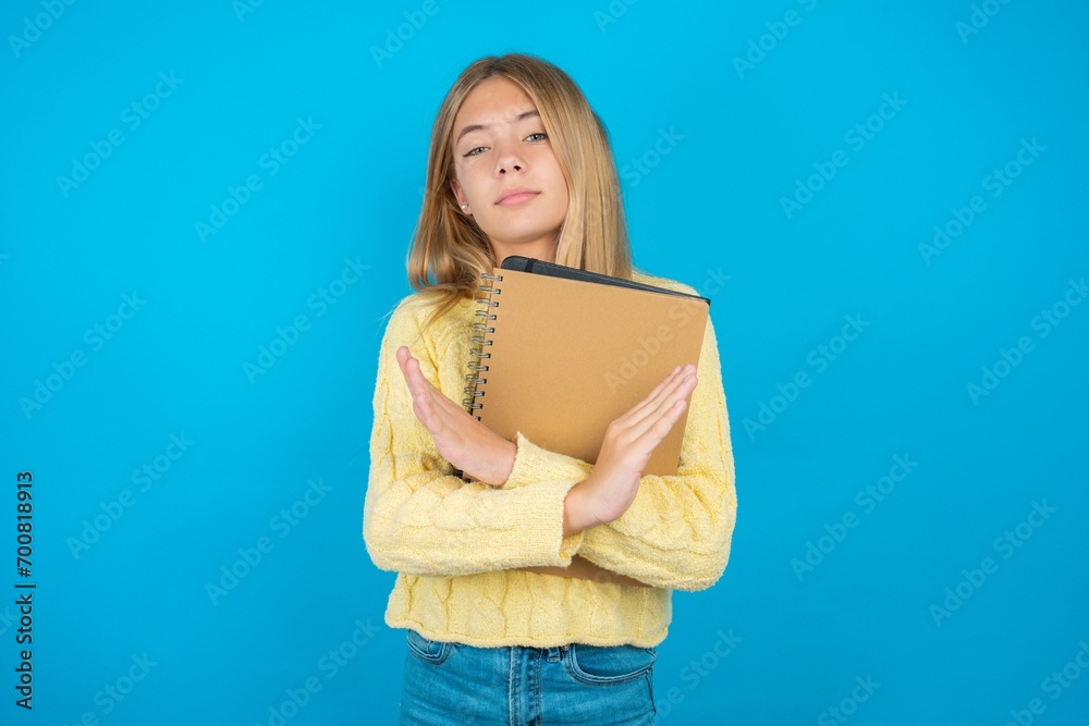 Beautiful kid girl wearing yellow sweater holding notebook Rejection expression crossing arms doing negative sign, angry face Beautiful kid girl wearing yellow sweater holding notebook Rejection expression crossing arms doing negative sign, angry face