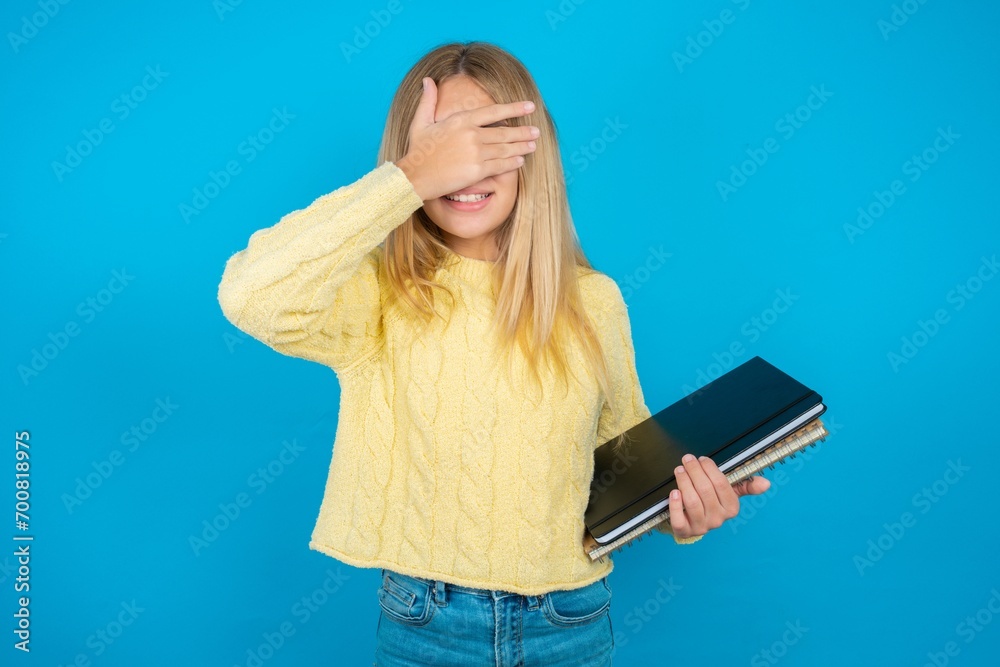 Beautiful kid girl wearing yellow sweater holding notebook smiling and laughing with hand on face covering eyes for surprise. Blind concept. Beautiful kid girl wearing yellow sweater holding notebook smiling and laughing with hand on face covering eyes for surprise. Blind concept.
