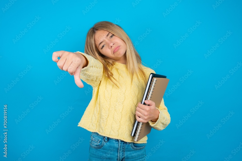Naklejka premium Beautiful kid girl wearing yellow sweater holding notebook feeling angry, annoyed, disappointed or displeased, showing thumbs down with a serious look