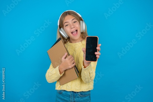 Beautiful kid girl wearing yellow sweater holding notebook with a mobile. presenting smartphone. Advertisement concept.