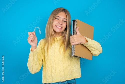 Beautiful kid girl wearing yellow sweater holding notebook holding an invisible braces aligner and rising thumb up, recommending this new treatment. Dental healthcare concept.