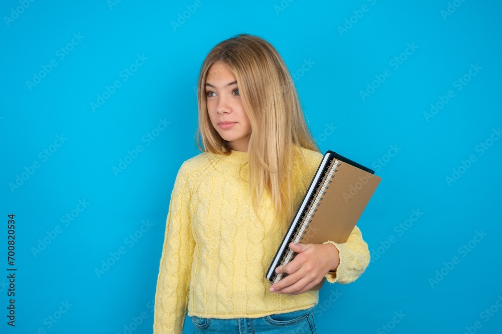 Beautiful kid girl wearing yellow sweater holding notebook stares aside with wondered expression has speechless expression. Embarrassed model looks in surprise Beautiful kid girl wearing yellow sweater holding notebook stares aside with wondered expression has speechless expression. Embarrassed model looks in surprise